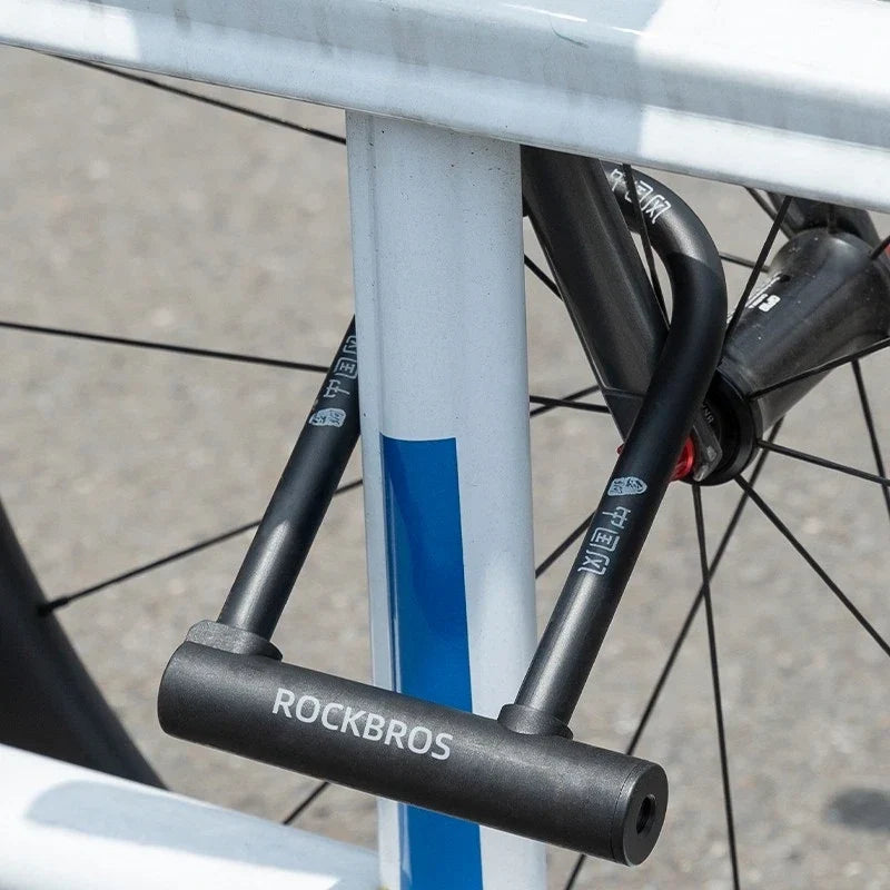 A close-up of a black Rockbros U-lock bicycle chain lock secured around a white and blue bicycle frame, with the bicycle wheel visible in the background.