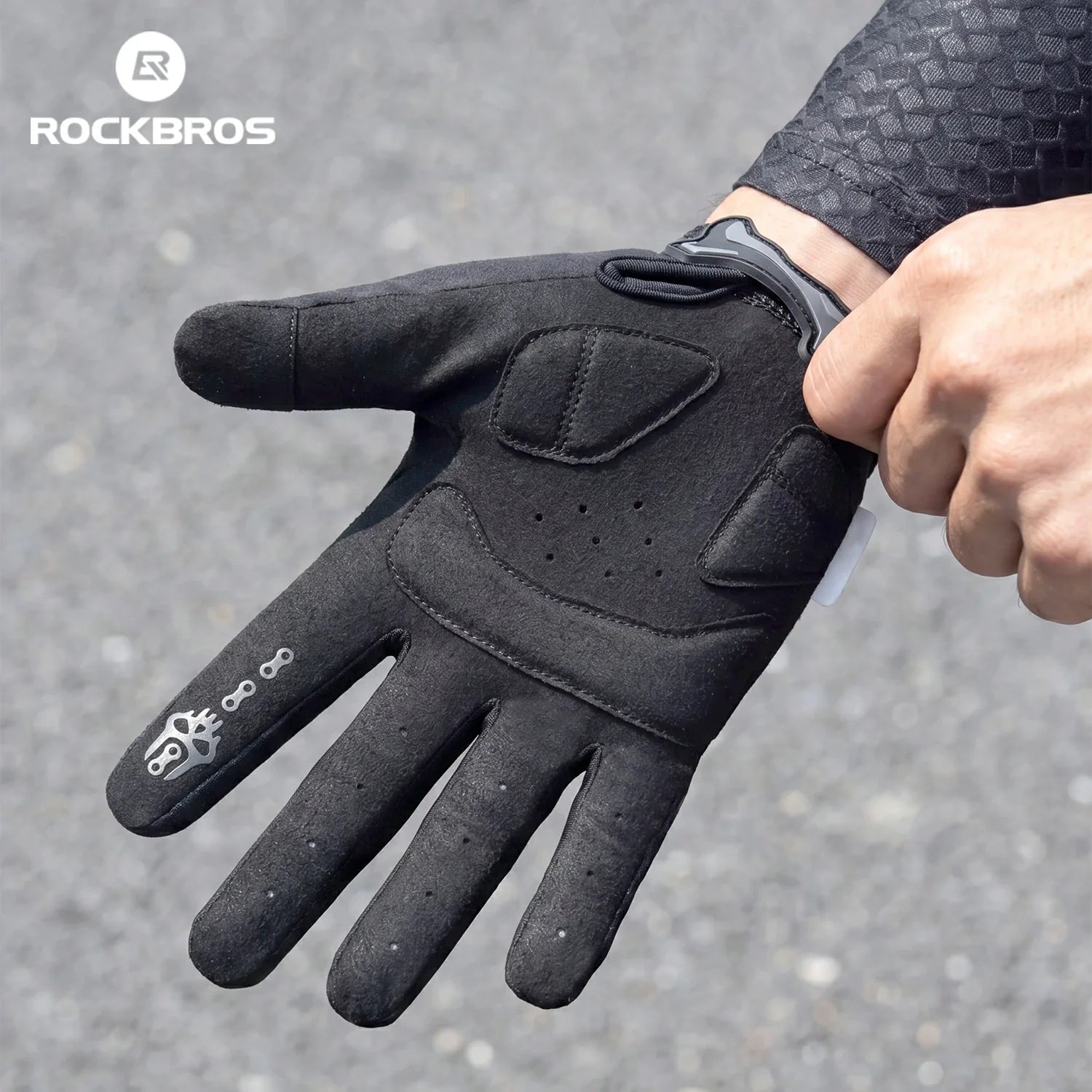 A black Rockbros cycling glove being worn on a hand, shown against a gray asphalt background with the Rockbros logo in the top left corner.