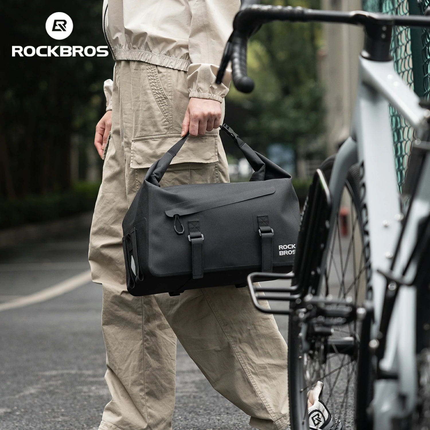 Person holding a black bag with 'RockBros' branding next to a bicycle on a street. #color_black