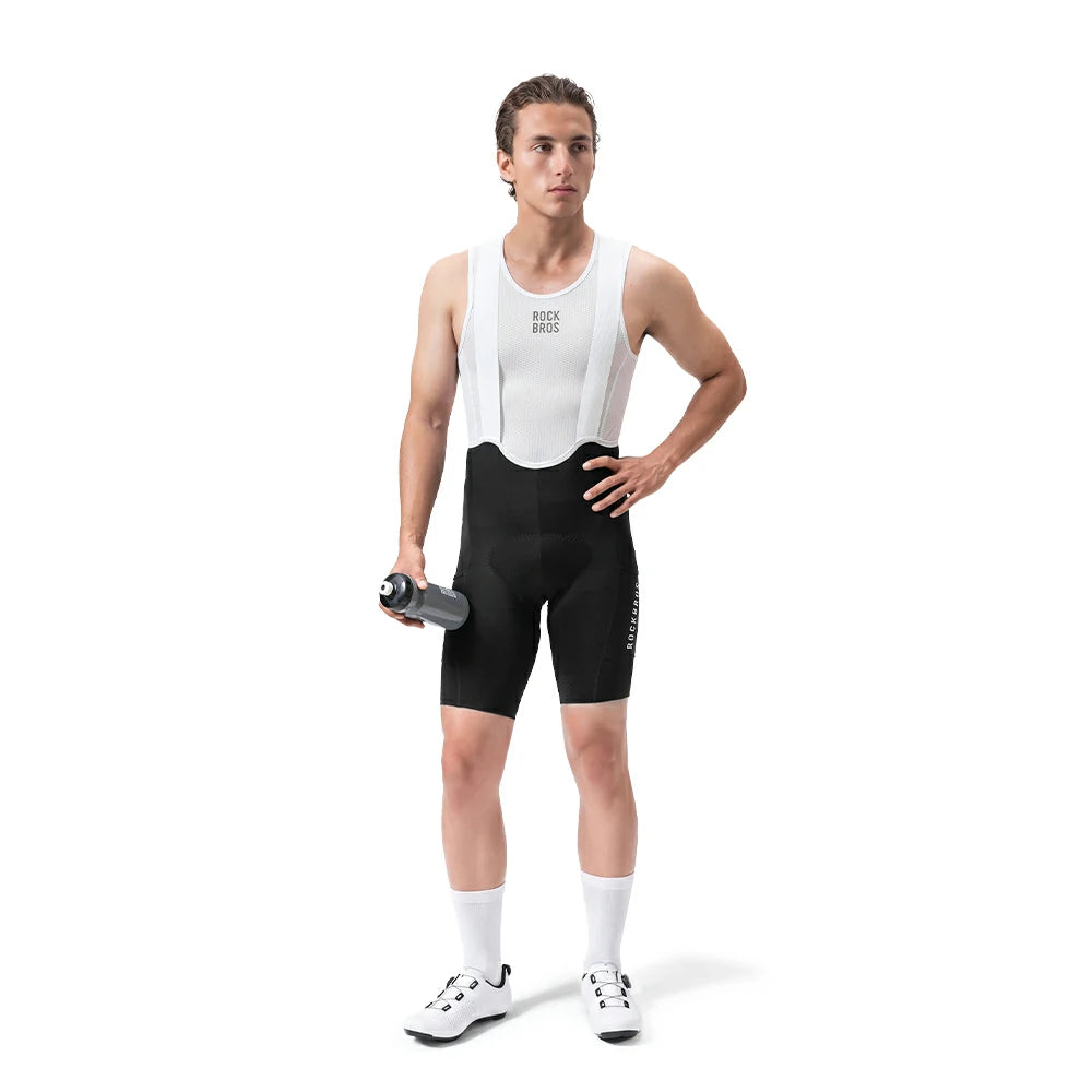 A male cyclist stands against a white studio background wearing a white bib shirt with black suspenders and black cycling shorts. He is holding a gray water bottle and wearing white socks and cycling shoes.