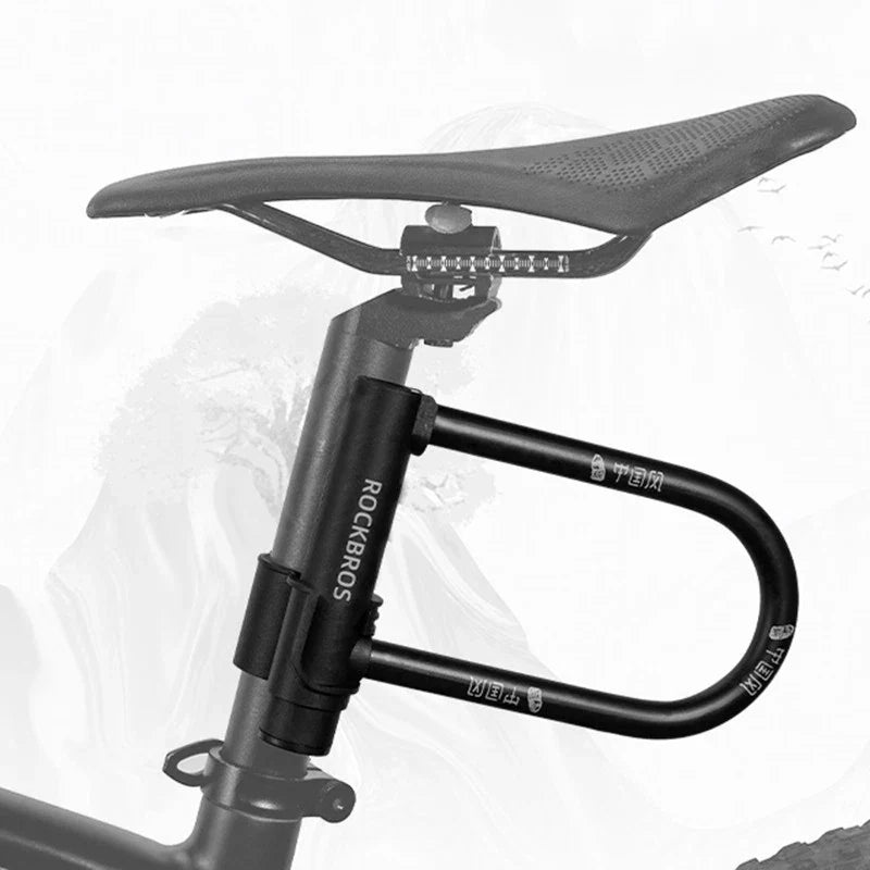 A black Rockbros bicycle seat post lock with a U-bar style lock, shown attached to a bicycle seat post with a saddle above, on a light gray background. 