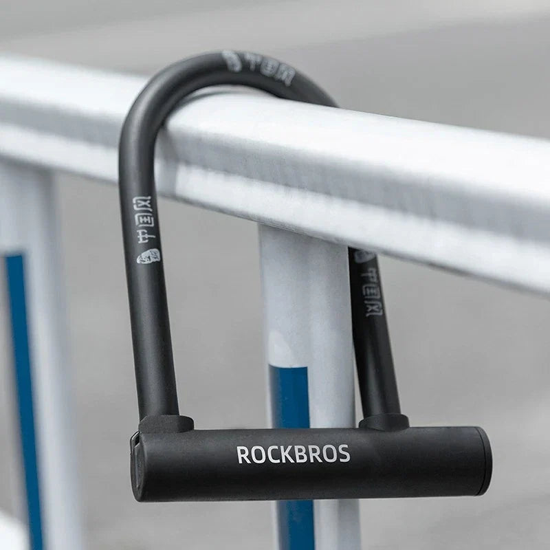 A black U-lock bicycle lock with the brand name ROCKBROS printed in white on the shackle, shown locked onto a white metal railing.