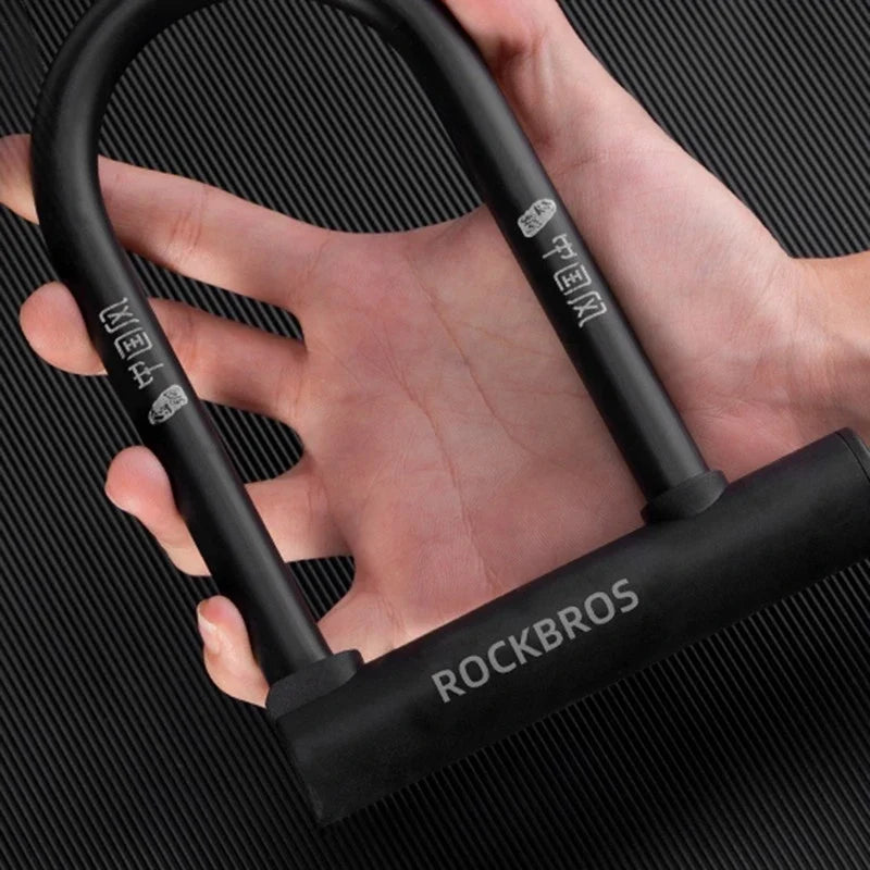 A close-up of a hand holding a black Rockbros U-lock bicycle lock against a dark textured background.