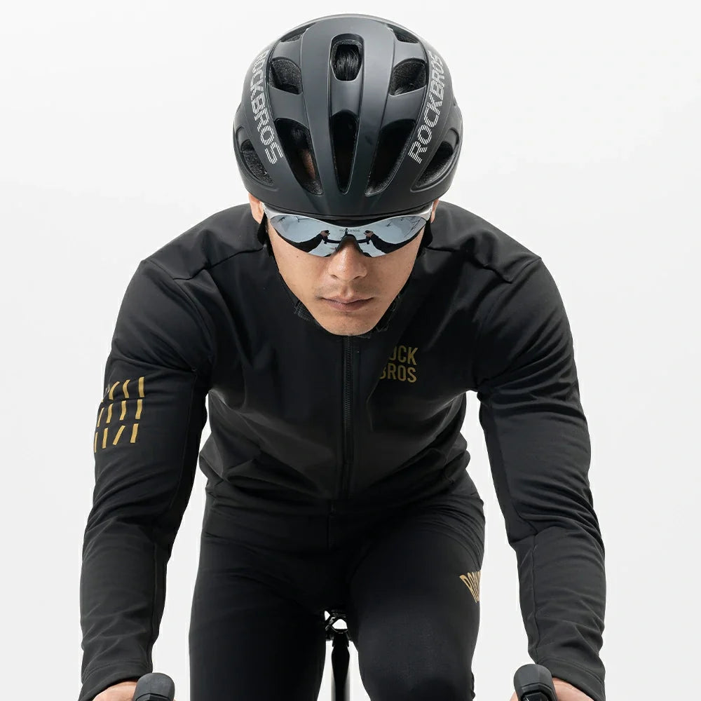 A cyclist in a black Rockros long-sleeve jersey and matching black cycling shorts, wearing a black Rockros helmet and sunglasses, captured from a front-facing angle against a clean white studio background.