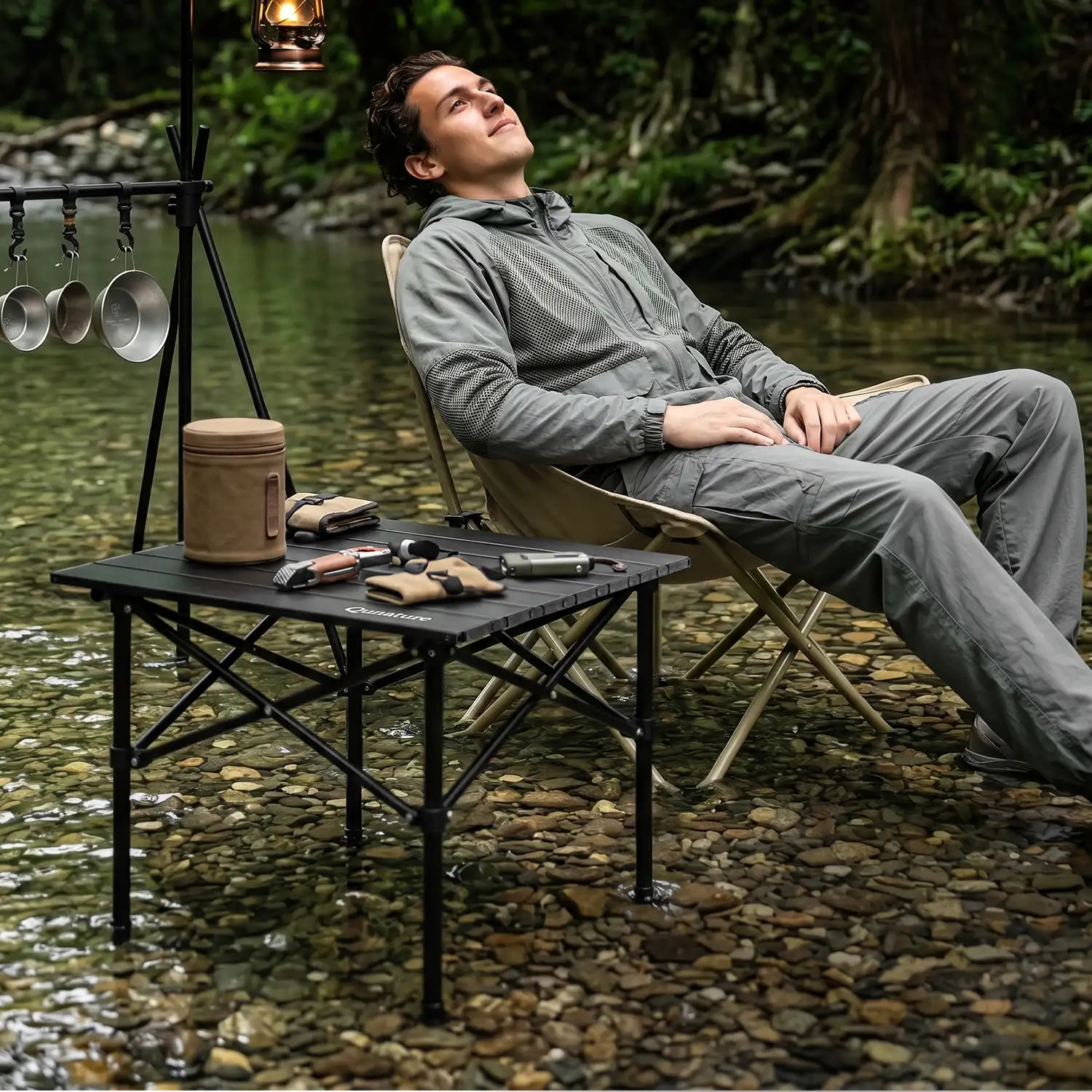 Man sitting by a river with camping gear, including a table and pots, in a natural setting.