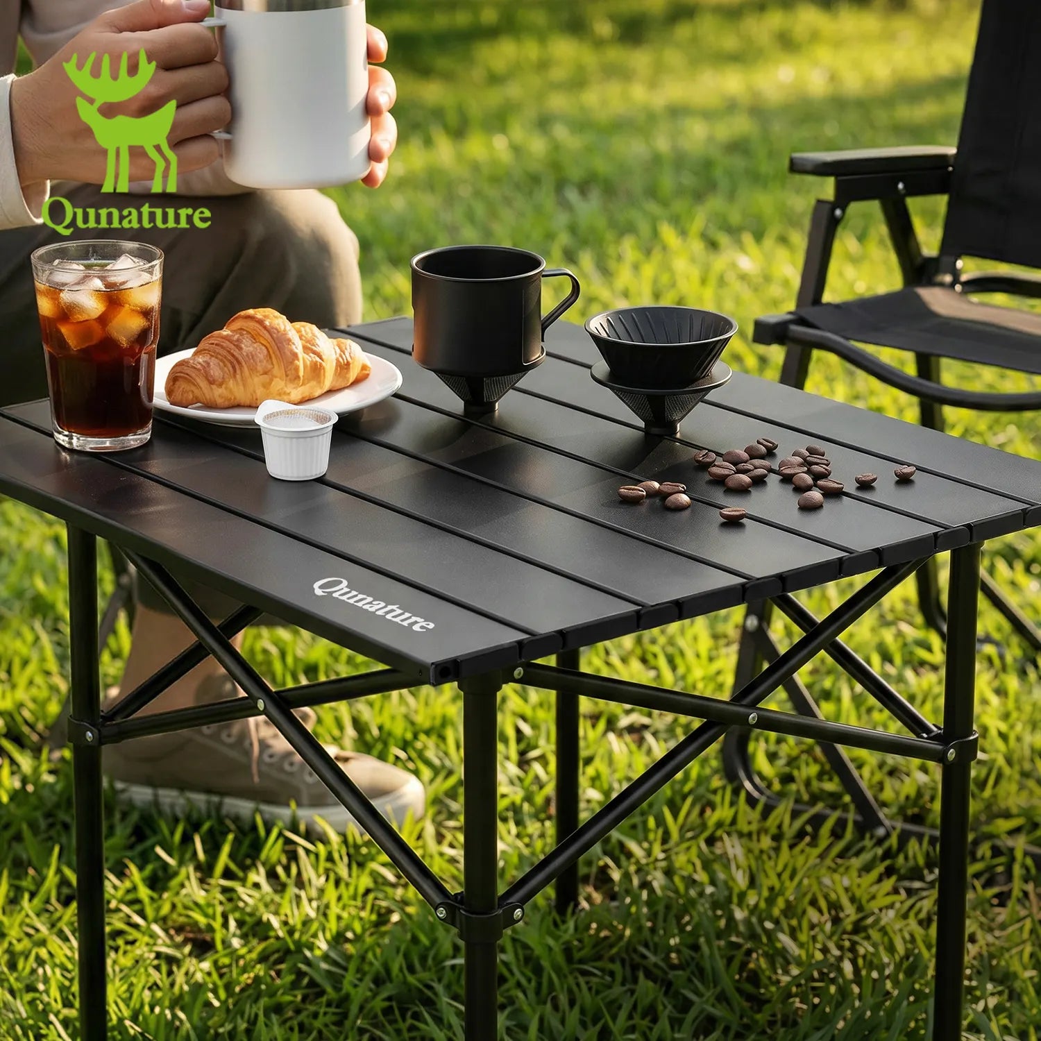 Person holding a mug with Qunature branding, sitting at a black camping table outdoors.
