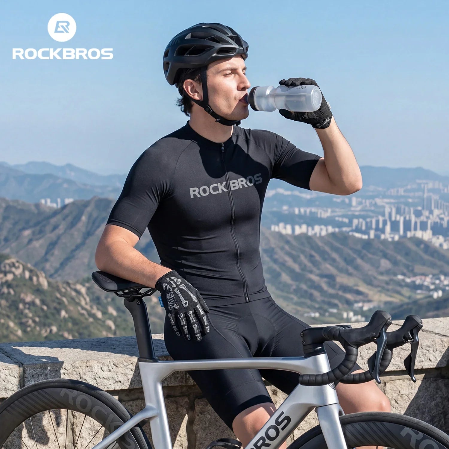 A man wearing a black ROCKBROS cycling jersey, matching shorts, a black helmet, and gloves sits on a silver road bike outdoors, drinking from a clear water bottle. The background shows a scenic mountain landscape under a blue sky, with the ROCKBROS logo in the top-left corner.