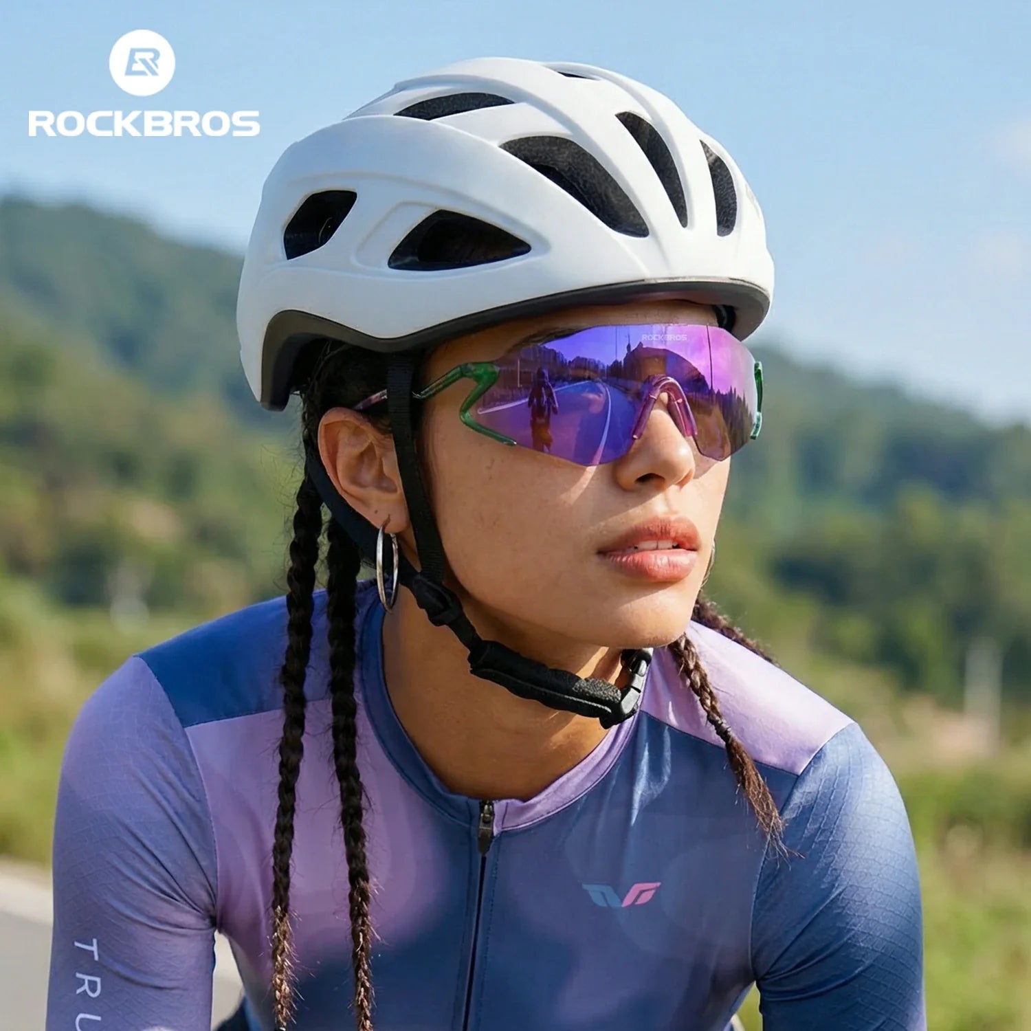 Person wearing a white helmet and purple cycling outfit with sunglasses, with mountains in the background. #Color_Purple