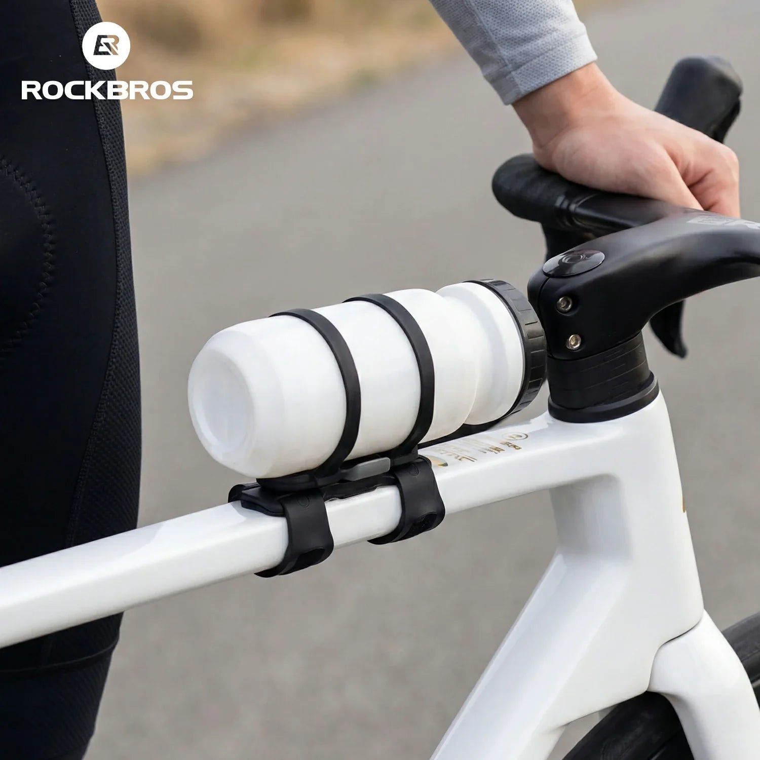 A close-up of a white water bottle mounted on a white bicycle frame with black straps, held by a rider whose hand and arm are visible, with the ROCKBROS logo in the top left corner.