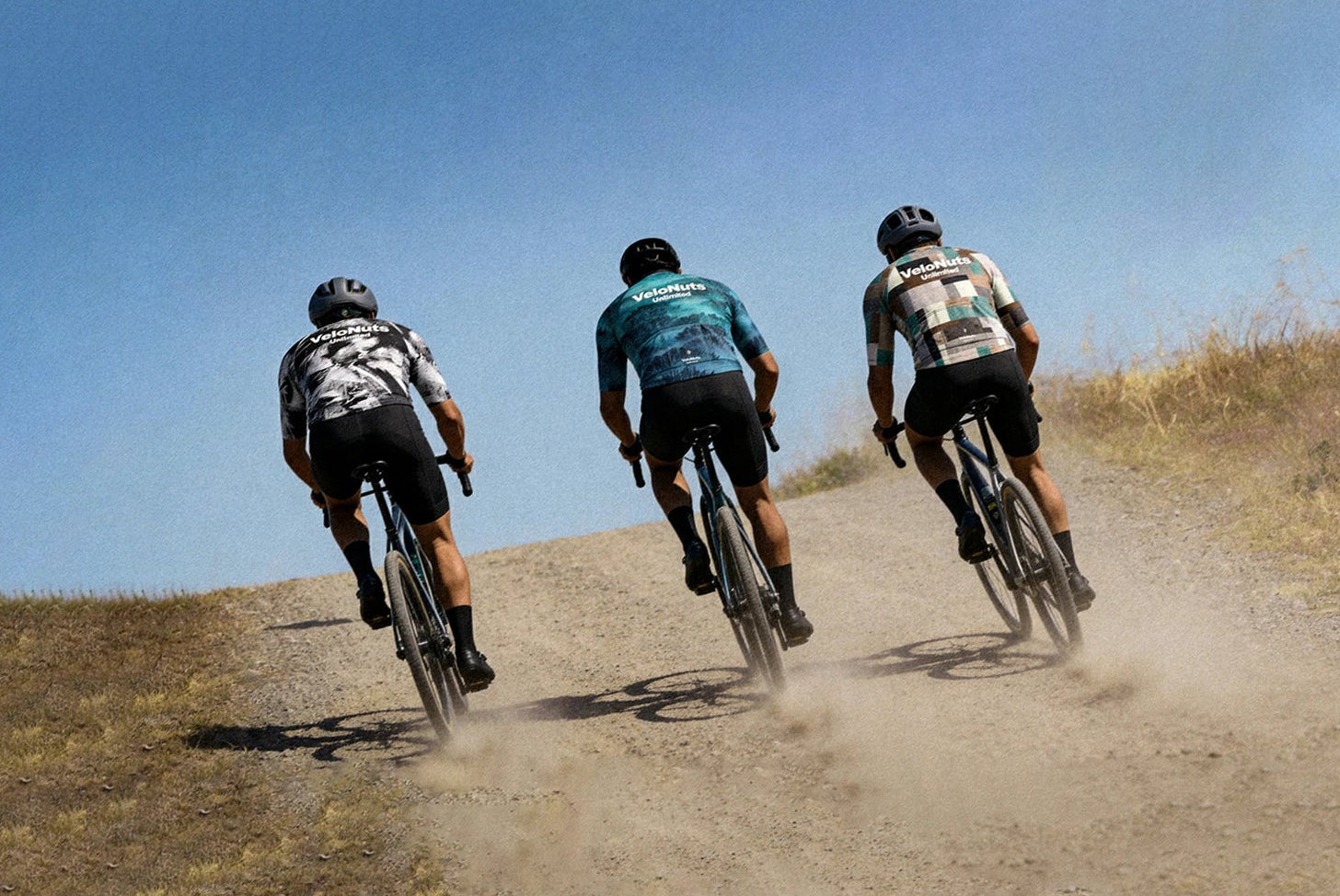 Three cyclists ride uphill on a dirt trail under a clear blue sky, kicking up dust. They wear cycling jerseys, shorts, and helmets, with dry grass and hills in the background.